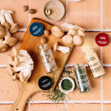 Mushrooms, nutmeg, and parsley on a wooden board with spice jars labeled Morton & Bassett, set on a tiled surface.