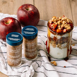 Yogurt parfait with granola and apples, next to ground cinnamon, nutmeg jars, and apples on a striped cloth.