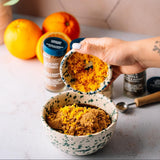 Hand adds orange zest to a bowl of brown sugar. Spices and fresh oranges are in the background on a kitchen counter.