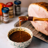 A close-up of a hand holding a basting brush dipped in a brown glaze, with a drip of the sauce falling into a speckled bowl. A large cooked ham is partially visible in the background on a metal rack, with blurred spice jars on a countertop.