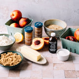 A styled flat-lay of baking ingredients on a tiled surface, including two jars of Ground Vanilla Extract, a bottle of M&R pure vanilla extract, flour, brown sugar, oats, butter, and fresh peaches and a lemon.