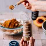 A hand holds a glass jar of Morton & Bassett Ground Nutmeg while another hand sprinkles the spice from a wooden spoon. A glass bowl filled with dry ingredients and sliced peaches are visible in the softly blurred background.
