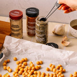 Hand scoops oregano from a jar near onion powder, harissa, garlic, and chickpeas on parchment, ready for seasoning.