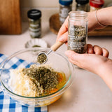 Hands sprinkle oregano into a bowl of ingredients on a blue checkered cloth, preparing a dish with spices and herbs.