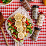 Grilled chicken with lemon slices, cucumber, and tomato on a fish-shaped plate, surrounded by spices on a checkered tablecloth.