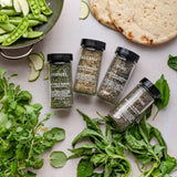 Jars of parsley, oregano, basil, and thyme with fresh greens, snap peas, and flatbread on a light surface.