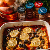 A white rectangular baking dish filled with baked fish topped with lemon slices, olives, and a red sauce, with several spice jars arranged behind it on a rustic wooden surface.