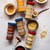 An overhead flat-lay of six jars of Morton & Bassett spices, including Turmeric, Ground Coriander, Paprika, Caram Masala, Ground Ginger, and a small bowl of whole spices, arranged on a light pink textured surface with small spoons and bowls of loose spices.