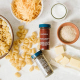 A styled flat-lay of pasta-making ingredients on a light stone surface, including shell pasta, shredded cheddar cheese, a bowl of milk, a terracotta bowl of flour, butter cubes, and two spice jars labeled Paprika and One True Love Italian Seasoning.