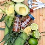 Two jars of Morton & Bassett San Francisco chili powder and Paprika seasoning arranged on a wooden cutting board with fresh ingredients including corn, avocado, lime, and green onions.
