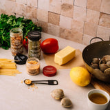 A styled kitchen countertop scene with food ingredients and cookware: a jar of dried herbs, a jar of pasta seasoning, a block of butter, a lemon, clams, spaghetti, parsley, a tomato, a metal pan with clams, and a small cup of tea, arranged against a beige tiled backsplash.