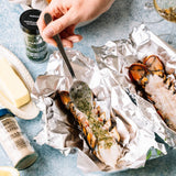 A close-up food scene showing a lobster tail topped with herb butter on aluminum foil, with a hand using a spoon to serve. Surrounding props include a bottle of Paskelly's herb butter, a bottle of Morton & Co salt, a pat of butter, and a glass of white wine on a blue textured surface.
