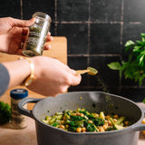 A person seasoning a dark gray pot of chickpeas and green vegetables with a wooden spoon, holding a small glass bottle of Oishi Turmeric and Ginger Spice powder in a kitchen setting.