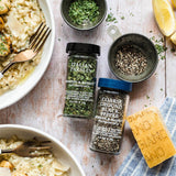 Two spice jars from Morton & Co. are displayed on a rustic white wooden table surrounded by food props, including a bowl of creamy pasta, a lemon wedge, a block of Parmesan cheese, and bowls of fresh parsley and ground black pepper.