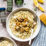 A top-down styled food scene showing a white bowl of creamy risotto with scallops, herbs, and shaved cheese, alongside bottles of coarse ground black pepper and Italian parsley, lemon wedges, a lemon wedge, and a blue striped cloth on a light wood surface.