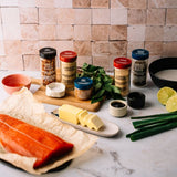 A styled kitchen scene with a salmon fillet on parchment paper, butter on a plate, herbs, lime, scallions, and several spice jars arranged on a marble countertop against a tiled backsplash.