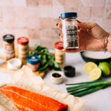 A hand holds a clear spice jar labeled Coast to Coast Black Pepper, with a blurred kitchen scene behind it including salmon, herbs, limes, green onions, and other seasoning jars.