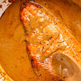 A close-up of a cooked salmon fillet in a creamy orange-brown sauce with visible black pepper, resting in a pan with a wooden spoon visible at the bottom right.