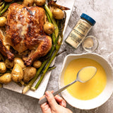 Overhead food scene showing a roasted whole chicken with potatoes and asparagus on a white platter, next to a bowl of yellow broth with a spoon and a bottle of ground white pepper.
