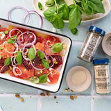 A styled flat-lay of a white rectangular baking dish filled with sliced blood oranges, red onion, and fresh basil leaves, surrounded by jars of Morton & Banquet Ground White Pepper and sea salt, with additional herbs and bowls on a light blue surface.