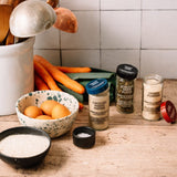 A styled kitchen countertop scene with several spice jars and bowls of ingredients, including ground white pepper, Italian herb blend, onion powder, coarse sea salt, flour, eggs, carrots, and a sweet potato, set against a tiled wall.