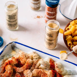 Seasoned shrimp in a baking dish surrounded by spice jars, including ground white pepper, garlic powder, and paprika, with a bowl of penne pasta on a white table.