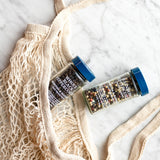 Two spice jars with blue lids labeled 'Whole Black Pepper' and 'Multicolored Pepper' arranged on a cream mesh bag over a white marble surface.