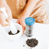 A person in a tan shirt uses a white mortar and pestle with whole black peppercorns, next to a glass jar of Shakerstyle Co. whole black peppercorns and a small dish of peppercorns on a white surface.