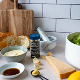 A jar of Whole Black Supercorns gourmet bresaol sauce and mustard, styled on a kitchen counter with bread, butter, cheese, and salad against a white subway tile background.