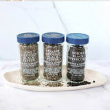 Three glass spice jars with blue lids labeled Morton & Basset black pepper varieties, arranged on a white tray against a light marble background.