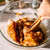 Marinated chicken pieces in a glass bowl with a wooden spoon, with a hand holding the spoon in a kitchen setting.