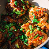 Close-up of glazed chicken pieces in a pan, topped with chopped green onions and sesame seeds.