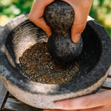 A close-up of a hand using a dark gray stone mortar and pestle to grind herbs or spices inside a textured stone bowl, set on a wooden surface with a blurred green outdoor background.