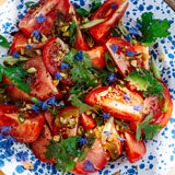 A close-up overhead shot of a fresh tomato salad in a white ceramic bowl with blue speckles. The salad features sliced red tomatoes, chopped green herbs, and small blue flowers, topped with a dressing.