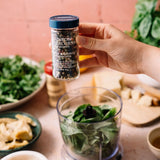 A hand holds a clear spice jar labeled Rainbow Percorns Sea Salt & Pepper Flakes above a blender filled with green leaves, with food ingredients and a pink background.