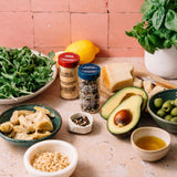 A styled food and spice arrangement on a stone surface against a terracotta tiled wall, featuring bowls of greens, artichokes, olives, cheese, avocado, lemon, and olive oil, with two spice jars centered and a basil plant in the background.