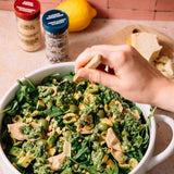 A close-up of a hand serving fresh green pesto from a white bowl filled with arugula, artichokes, and olives. Two spice jars and a lemon sit in the background on a kitchen counter.