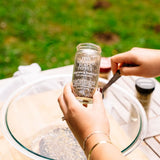A person holding a glass jar of Fine Organics Black Pepper over a large glass bowl outdoors, with grass visible in the blurred background.