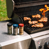 Three spice seasoning bottles labeled Pepper Jack, Garlic Pepper, and Garlic Powder sit on the side of a stainless steel grill with chicken cooking on the grate and flames visible.