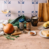 Two jars of gourmet spices displayed on a rustic wooden table with cooking ingredients including an onion, garlic, fresh rosemary, a glass of white wine, and cheese, set against a tiled wall.