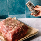 A hand with dark nail polish sprinkles ground black pepper from a clear shaker onto a raw beef roast resting on parchment paper in a baking pan, set against a blue tiled background.