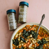 Bowl of soup with vegetables and herbs, accompanied by jars of black pepper and oregano on a pink background.