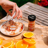 A hand pours liquid from a glass into a plate of seasoning with visible cayenne pepper flakes, next to a bottle of Cayenne Pepper seasoning on a wooden table with orange slices.