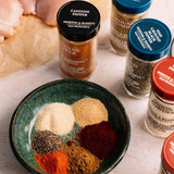 A bowl filled with six different ground spice powders in various colors, surrounded by several jars of Morton & Bassett spices including cayenne pepper, ground cumin, ground ginger, and onion & garlic powder, with raw chicken pieces in the background.