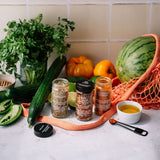 Three glass spice jars from The Spice Shop are arranged on a light stone surface with fresh ingredients around them, including a cucumber, parsley, a watermelon in an orange mesh bag, sliced peppers, a small bowl of yellow dressing, and a spoon.