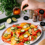 A styled food scene on a light marble surface with a large white platter filled with sliced watermelon, yellow and orange heirloom tomatoes, cucumber rounds, jalapeño slices, and chopped cilantro. A hand is sprinkling herbs over the salad.