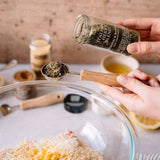 A person's hands holding a glass jar labeled 'Italian Seasoning Blend' over a measuring spoon filled with the dried herb blend, with a glass bowl and other kitchen items in the background.
