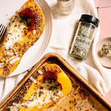 A styled overhead food scene showing baked stuffed squash halves with melted cheese and herbs, served on a white plate and in a baking pan, alongside a spice jar labeled Italian Seasoning Blend and a small dish of dried herbs on a cream cloth.