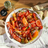 A white bowl filled with a tomato and vegetable salad, including sliced red tomatoes, orange carrots, red onions, and green herbs, set on a wooden table with a spoon, napkin, and small jar of dried herbs.