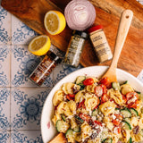 A styled overhead shot of a prepared pasta salad in a white bowl with wooden spoons, surrounded by seasoning bottles, a lemon, a sliced red onion, and a wooden cutting board on a blue-and-white patterned tile surface.